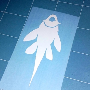 May include: A white vinyl decal of a stylized creature, possibly a fish or a mythical being, with a rounded head and elongated body. The decal is on a light blue background, possibly a cutting mat, with grid lines.