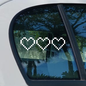 Puede incluir: Adhesivo blanco de corazón pixelado aplicado a una ventana de coche. El diseño presenta tres corazones conectados, creando un diseño simple pero llamativo. La ventana del coche refleja el entorno.