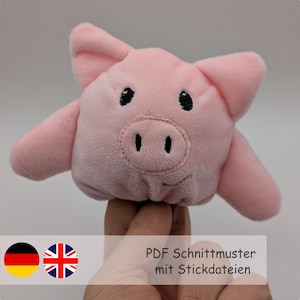 May include: Pink plush pig finger puppet with black eyes and a black snout. The puppet is being held by a hand. The text "PDF Schnittmuster mit Stickdateien" is visible below the puppet.