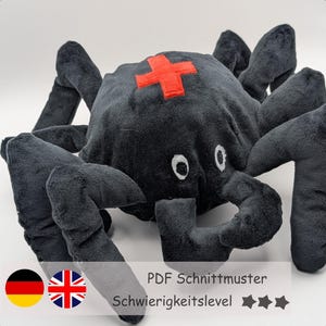 Puede incluir: Un peluche negro con forma de araña, con una cruz roja en la cabeza y ocho patas. La araña tiene ojos blancos y una boca curvada. El texto en la imagen dice "PDF Schnittmuster" y "Schwierigkeitslevel".