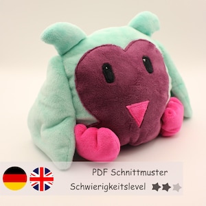 May include: A plush toy shaped like an owl with a heart-shaped face. The owl is green, pink, and purple. The toy is sitting on a white surface. PDF Schnittmuster Schwierigkeitslevel ***
