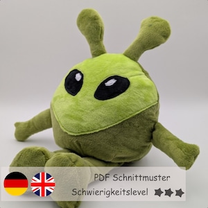 Op de afbeelding: Een groene pluche alien speelgoed met grote zwarte ogen en antennes. Het zachte speelgoed heeft uitgestrekte armen en zit. De afbeelding bevat de tekst "PDF Schnittmuster" en "Schwierigkeitslevel".