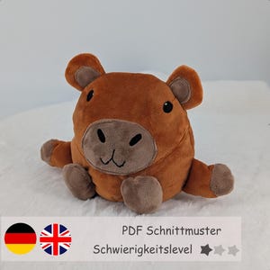 May include: A round, plush capybara toy with soft brown fur, a lighter brown muzzle, and small dark embroidered eyes, nose, and mouth. The toy features two small ears, two front paws, and two back feet, all in a matching light brown fabric.