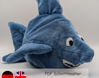 May include: A blue plush shark toy with white eyes and teeth. The toy is lying on a white surface. The text "PDF Schnittmuster" and "Schwierigkeitslevel" is visible below the toy. Three stars are shown, two are black and one is gray.