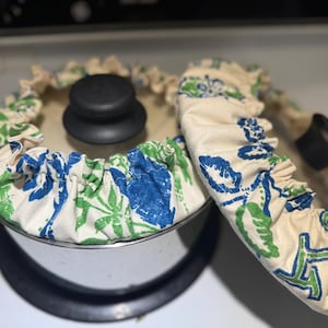 Op de afbeelding: Een roestvrijstalen pan met een zwarte deksel en een stoffen hoes met bloemenprint. De stof is wit met blauwe en groene bloemenmotieven.