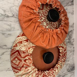 Op de afbeelding: Twee ronde dekselhoezen voor potten met een geometrisch patroon. De bovenste hoes is oranje met een wit geometrisch patroon. De onderste hoes is wit met een rood geometrisch patroon. Beide hoezen hebben een zwarte knop in het midden.