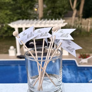 May include: A clear glass tumbler containing several wooden skewers, each topped with a small white paper flag. Each flag features the text The Mills printed in a black script font. The skewers have small wooden beads at the very top ends.