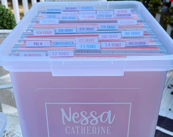 May include: A pink plastic storage bin with a clear lid, filled with labeled file folders. The labels include categories like "Newborn," "Kindergarten," and "Graduation." The bin is labeled "Nessa Catherine."