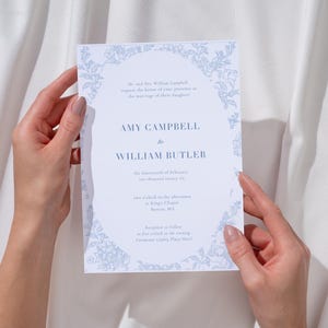 May include: A light blue and white wedding invitation with floral border, held by two hands. The invitation reads "Amy Campbell to William Butler" with event details. The background is a white fabric.