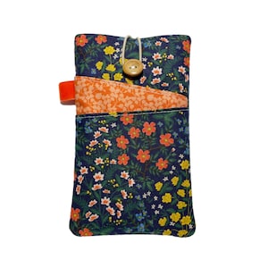 May include: A rectangular fabric pouch featuring a dark blue background with a multi-coloured floral print. It includes an orange patterned front pocket, a light-coloured elastic cord closure, and a single wooden button on the top flap.