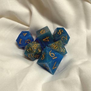 May include: A collection of seven polyhedral dice in shades of blue and teal with gold numbering. The dice are made of a translucent material, with swirls of color and gold flecks. The dice are for tabletop games.
