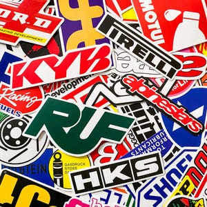 Pode incluir: Uma cole&ccedil;&atilde;o de autocolantes coloridos com temas de corridas e autom&oacute;veis. Os autocolantes apresentam v&aacute;rios logotipos e texto em vermelho, branco, preto e verde. Alguns autocolantes incluem as marcas KYB, Pirelli e HKS.