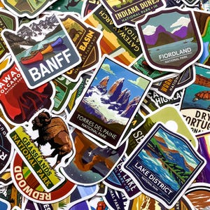 Puede incluir: Una colección de pegatinas coloridas con diseños de parques nacionales. Las pegatinas muestran varios paisajes, incluyendo montañas, lagos y vida silvestre. El texto incluye "Banff", "Torres Del Paine" y "Lake District".