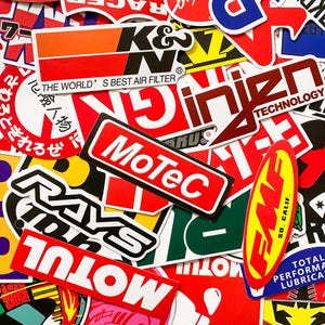Pode incluir: Uma cole&ccedil;&atilde;o de autocolantes coloridos com v&aacute;rios log&oacute;tipos e texto. Os autocolantes apresentam marcas como K&N, MoTeC e Rays, com designs em vermelho, branco, preto e amarelo. Alguns autocolantes t&ecirc;m texto japon&ecirc;s.