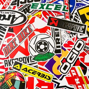 Pode incluir: Uma cole&ccedil;&atilde;o de autocolantes coloridos com v&aacute;rias marcas e log&oacute;tipos. Os autocolantes incluem "DIRT BIKE", "AKRAPOVIC", "OGIO" e "ACERBIS". A imagem &eacute; um close-up com v&aacute;rias formas e tamanhos.