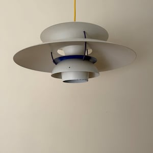 May include: A modern, multi-tiered pendant light fixture. The light features a large, circular shade in a light gray color, with a smaller, blue accent ring. A yellow cord suspends the light from the ceiling.