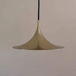 Puede incluir: Una lámpara colgante moderna con una pantalla metálica en forma de campana de color dorado claro. Cuelga de un cable negro. El diseño es minimalista y versátil.