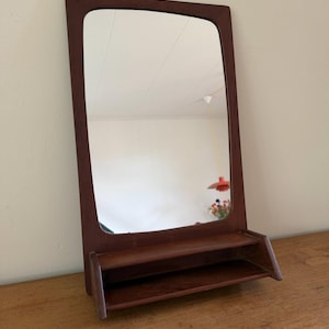 Op de afbeelding: Een rechthoekige wandspiegel met een donkerbruin houten frame en een ingebouwde horizontale plank aan de onderkant. De spiegel heeft afgeronde bovenhoeken en een gladde, gepolijste houten afwerking voor een helder reflecterend oppervlak.