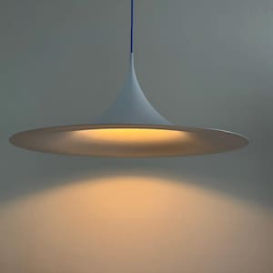 Puede incluir: Una lámpara colgante blanca con una pantalla ancha y acampanada en forma de platillo, suspendida por un cable azul fino. El interior de la pantalla emite un brillo dorado, cálido y suave, que ilumina el área bajo la lámpara en un círculo.