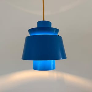 May include: A bright blue pendant light fixture with a tiered, multi-layered cylindrical design. The lamp features a smooth, glossy finish and is suspended from a thin, light-colored cord, emitting a soft glow from its central opening.