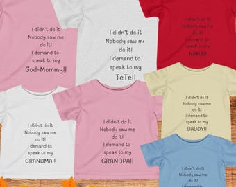 Op de afbeelding: Een set van zes roze, witte, rode, gele, blauwe en lichtblauwe t-shirts met een humoristische boodschap op elk. De boodschappen zijn: "Ik heb het niet gedaan, Niemand heeft me gezien! Ik eist om met mijn God-Mama te praten!"  "Ik heb het niet gedaan, Niemand heeft me gezien! Ik eist om met mijn TeTe te praten!"  "Ik heb het niet gedaan, Niemand heeft me gezien! Ik eist om met mijn Nana te praten!"  "Ik heb het niet gedaan, Niemand heeft me gezien! Ik eist om met mijn Oma te praten!"  "Ik heb het niet gedaan, Niemand heeft me gezien! Ik eist om met mijn Opa te praten!"  "Ik heb het niet gedaan, Niemand heeft me gezien! Ik eist om met mijn Papa te praten!"  "Ik heb het niet gedaan, Niemand heeft me gezien! Ik eist om met mijn Mama te praten!"