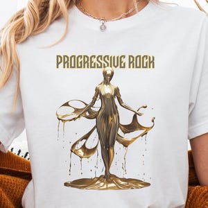 以下が含まれることがあります： 「PROGRESSIVE ROCK」の文字と、流れるようなドレープのある人物の金色のイラストが描かれた白いTシャツ。人物は金の台座の上に立っています。モデルはデニムショーツとマスタード色のカーディガンを着ています。