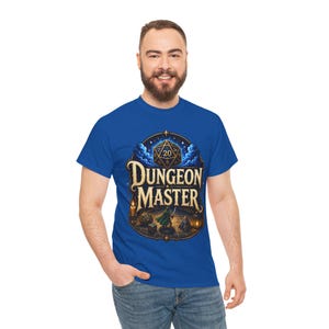 Puede incluir: Una camiseta azul real con un estampado gráfico que incluye el texto "DUNGEON MASTER" en una fuente serif dorada. El diseño presenta un dado de veinte caras grande, una figura pequeña con una espada, velas y otros dados poliédricos sobre un fondo de cielo estrellado azul oscuro dentro de un marco ovalado ornamentado.