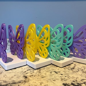 May include: A row of colorful butterfly-shaped decorations. The butterflies are in shades of purple, yellow, and teal, each mounted on a white rectangular base. The background is a light blue wall.