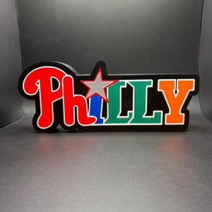 May include: A decorative sign featuring the word PHILLY in bold, multi-colored lettering. The P is red, the H is red, the I is replaced by a silver star over a blue vertical bar, the Ls are green, and the Y is orange, all on a black background.