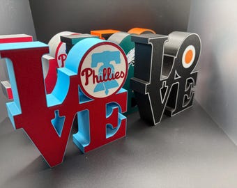 May include: A collection of three-dimensional block letters spelling LOVE, featuring sports team logos. The front letter is light blue and red with a Phillies logo. Another block is black with a white outline and an orange circle logo.