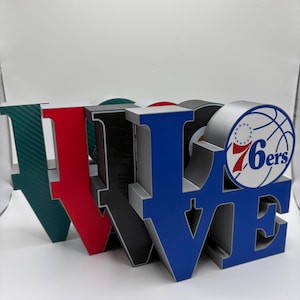 May include: A decorative sign spelling "LOVE" in large, block letters. The letters are in various colors: green with a carbon fiber pattern, red, black, and blue. The letter "E" features a basketball logo with "76ers" text.