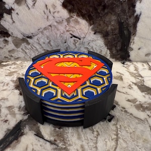 May include: A stack of round coasters featuring the Superman logo in red and yellow. The coasters have a blue and yellow honeycomb pattern and are held in a black holder. The Superman logo is prominently displayed on the top coaster.