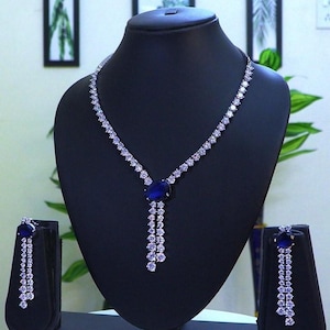 Può includere: Un set di collana e orecchini argentati esposto su un busto di gioielli nero. La collana presenta un grande pendente ovale di pietra preziosa blu scuro con un design a cascata di pietre preziose più piccole e trasparenti. Gli orecchini rispecchiano il design del pendente.