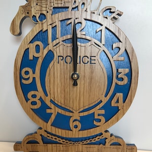 May include: A wooden clock with a blue face and a police badge design. The clock has a black hand and a gold second hand. The clock is shaped like a police badge and has a gun and handcuffs on the side. The clock says "Police" on the badge.