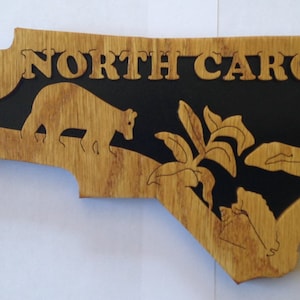May include: A wooden cutout of the state of North Carolina with a black background. The state name is written in white letters across the top. The cutout features a bear, a plant, and a woman sitting on a log.