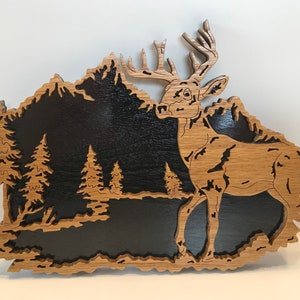 May include: A wooden cutout of a deer standing in a forest scene. The deer is brown and has large antlers. The forest is black and has trees and mountains. The cutout is made of wood and has a natural finish.