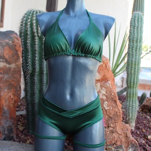 May include: A dark green two-piece bikini set featuring a triangle top with thin halter straps and matching low-rise bottoms with a V-shaped waistband. The set includes two thin leg bands that wrap around the upper thighs.
