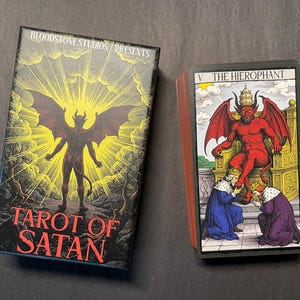 May include: A Tarot of Satan deck by Bloodstone Studios featuring a box with a winged figure against a yellow sunburst and a card showing The Hierophant with a red horned figure seated on a throne while two figures kneel before it.