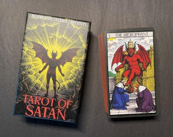 May include: A Tarot of Satan deck by Bloodstone Studios featuring a box with a winged figure against a yellow sunburst and a card showing The Hierophant with a red horned figure seated on a throne while two figures kneel before it.