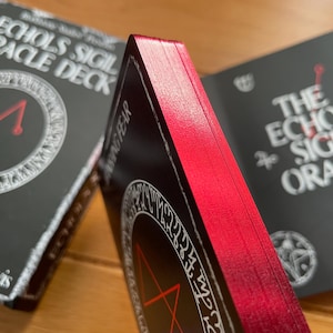 May include: Two decks of oracle cards with black backgrounds and red edges. The top deck has the text "Echoes Sigil Oracle Deck" and a white circle with a red triangle inside. The bottom deck has the text "The Echoes Sigil Oracle" and a white circle with a red pentagram inside.