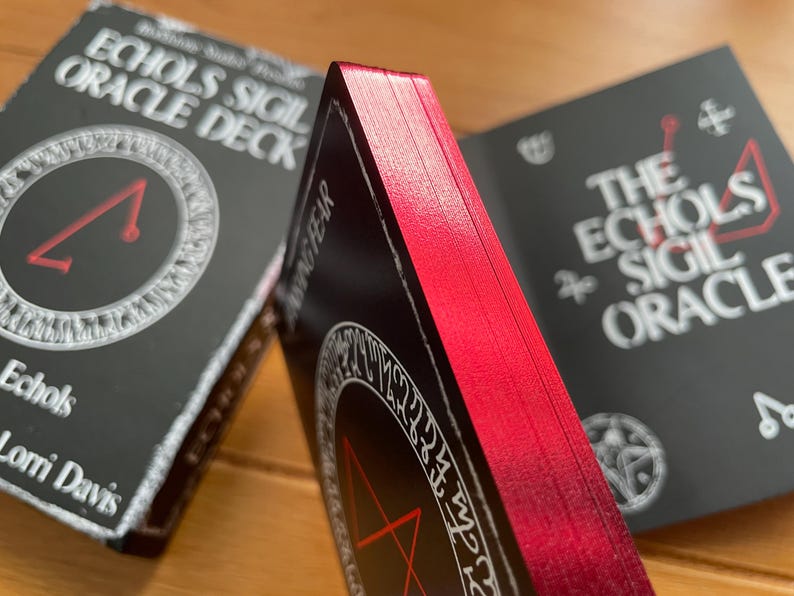 May include: Two decks of oracle cards with black backgrounds and red edges. The top deck has the text "Echoes Sigil Oracle Deck" and a white circle with a red triangle inside. The bottom deck has the text "The Echoes Sigil Oracle" and a white circle with a red pentagram inside.