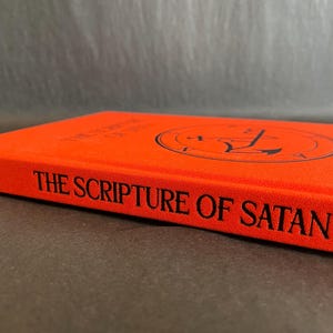 May include: A bright orange hardcover book with the title THE SCRIPTURE OF SATAN printed in black on the spine. The front cover features a circular black sigil design centered on the orange fabric material, showcasing a clean, minimalist aesthetic.