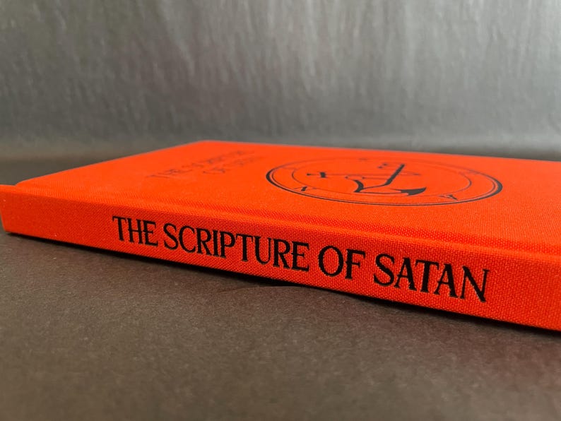 May include: A bright orange hardcover book with the title THE SCRIPTURE OF SATAN printed in black on the spine. The front cover features a circular black sigil design centered on the orange fabric material, showcasing a clean, minimalist aesthetic.