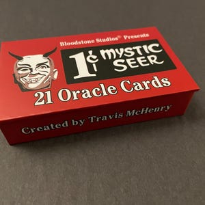 May include: A rectangular red box for 21 Oracle Cards titled 1¢ Mystic Seer. The box features a white illustration of a horned face on the left, a black rectangular logo with white text, and the creator name Travis McHenry printed in white.