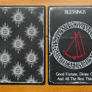 May include: A black and white tarot card with the word "Blessings" at the top. The card features a red symbol inside a circle with white text around the outside. The back of the card has a repeating pattern of white eyes inside a circle with white rays.