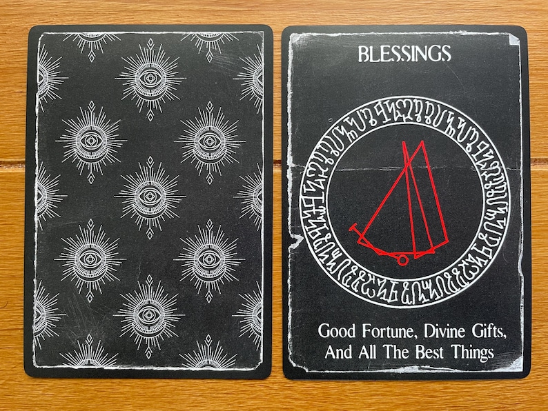 May include: A black and white tarot card with the word "Blessings" at the top. The card features a red symbol inside a circle with white text around the outside. The back of the card has a repeating pattern of white eyes inside a circle with white rays.