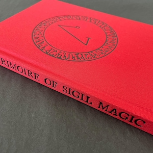 May include: A red book titled "Grimoire of Sigil Magic" with a black circular design on the cover. The book's spine displays the title in black lettering. The cover has a textured appearance.