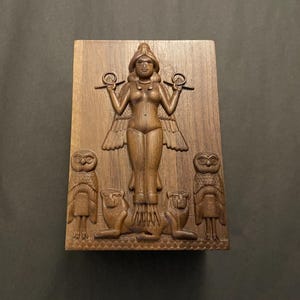 Puede incluir: Una talla de madera rectangular que presenta una figura femenina central con alas, sosteniendo objetos. Dos búhos flanquean la figura, con dos criaturas parecidas a leones debajo. La talla es en tonos marrones.