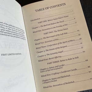 May include: An open book displaying a Table of Contents page with black text on a cream-colored background. The text lists sections including Part One: Before Satan Had A Name, Part Two: The Divine Satan, and Part Three: Better to Rule in Hell.