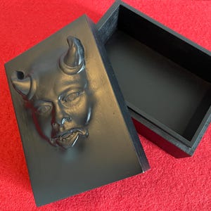 May include: A rectangular black wooden box with a detachable lid. The lid features a three-dimensional molded relief of a face with two curved horns protruding from the top. The interior of the box is hollow and finished in a matching matte black.